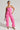 A woman exudes confidence wearing the Bubblegum Shorty Corset Top in Pink, paired with matching pants. Her pink faux leather outfit is accessorized with sunglasses, hoop earrings, and a small patterned bag. She strikes a stylish pose in open-toed heels against a plain backdrop.