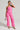 A person stands confidently in the vibrant Bubblegum Shorty Corset Top in Pink, paired with a sleek jumpsuit featuring multiple pockets. They hold a stylish white handbag and accessorize with sunglasses, hoop earrings, and strappy high heels. The plain background accentuates their bold ensemble.