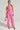 A woman in the BUBBLEGUM SHORTY CARGO PANT IN PINK stands confidently against a plain background. She holds a small white handbag with pink accents and 'Paris' written on it. She wears hoop earrings, sunglasses, and strappy high-heeled sandals that complement her bright pink faux leather outfit.