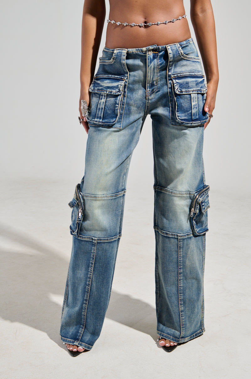 Dressed in the LOW RISE SHORTY OMBRE CARGO DENIM, which showcases a faded, distressed style, an individual stands with their hands in the multiple pockets of these wide-leg jeans. The Y2K aesthetic is highlighted by a stylish belt and jewelry adorning one hand.