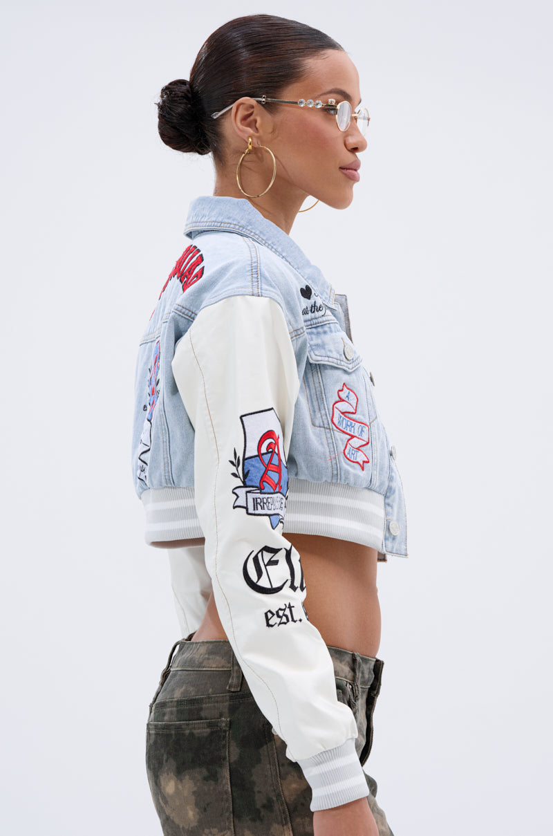 A woman wearing glasses and hoop earrings models the OFF CAMPUS DENIM CROP BOMBER with embroidered patches and faux leather sleeves, paired with camo-print pants, standing in profile against a plain light background.