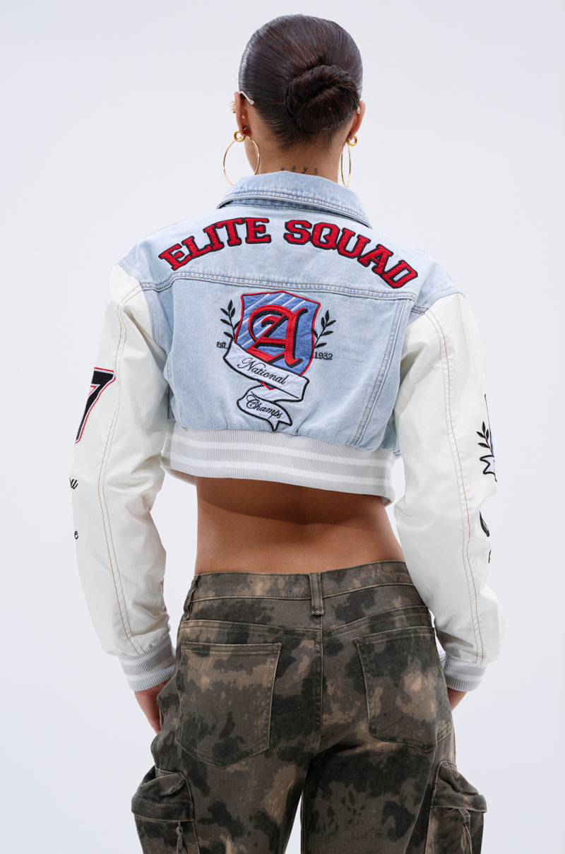 A woman with her hair in a bun wears the OFF CAMPUS DENIM CROP BOMBER, a light blue and white varsity-style jacket with faux leather sleeves and "Elite Squad" patches on the back, paired with camo cargo pants. She faces away from the camera.