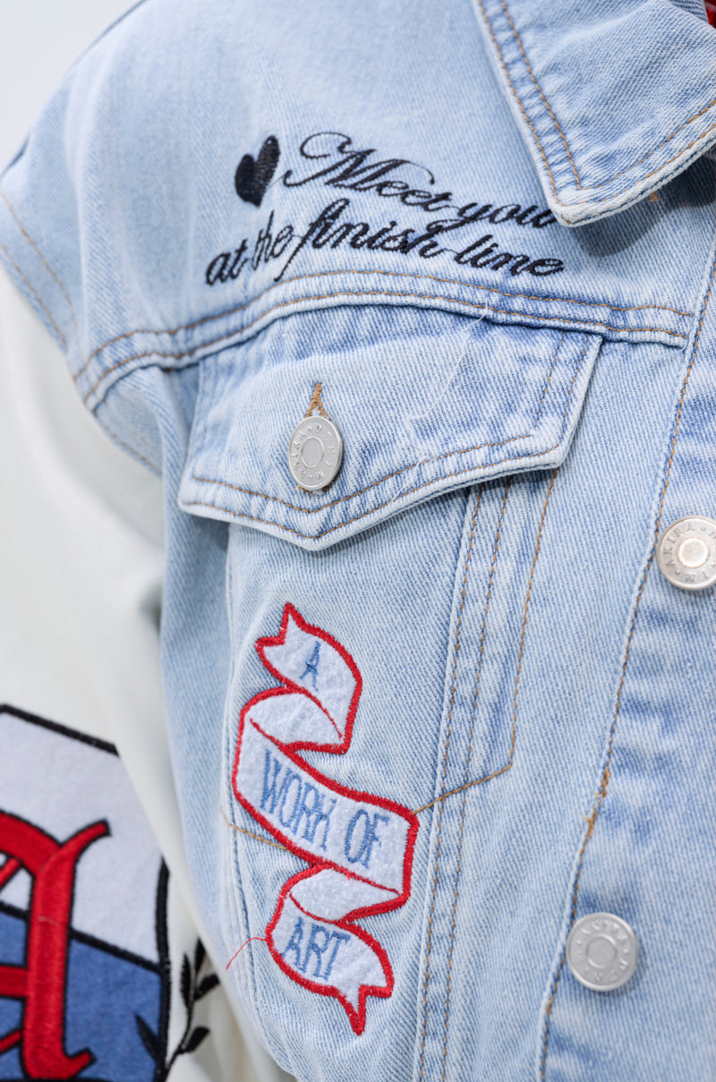 Close-up of the OFF CAMPUS DENIM CROP BOMBER in light blue, featuring embroidered text. A pocket ribbon reads "A WORK OF ART" in red and white; above it, black script says "See you at the finish line" with a black heart.