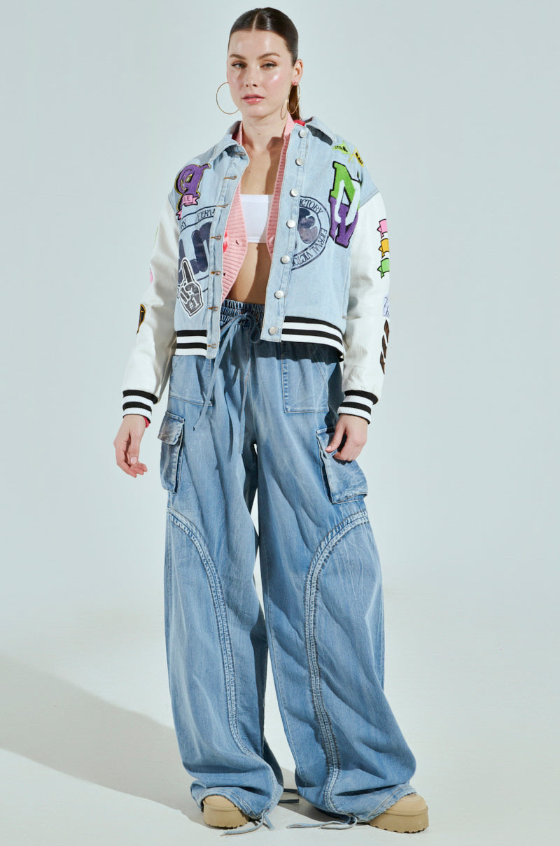 A person wears the ACADEMIA DENIM BOMBER, featuring a colorful mixed media design, a white crop top, and baggy light blue jeans. They have light brown hair tied back, large hoop earrings, beige shoes, and stand against a plain background.