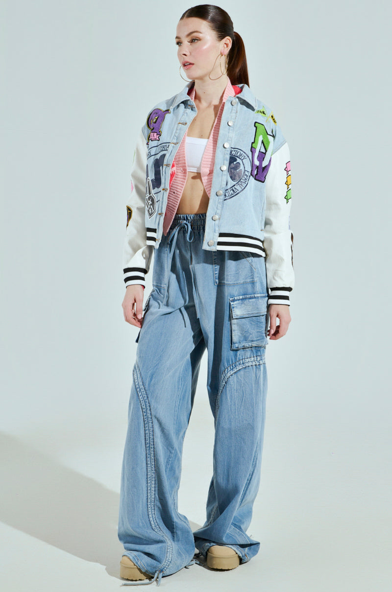 A woman poses against a light backdrop in the ACADEMIA DENIM BOMBER, featuring colorful patches. She complements it with a white crop top, loose blue denim pants, and beige shoes. Her hair is neatly tied back as she stands with one hand by her side.