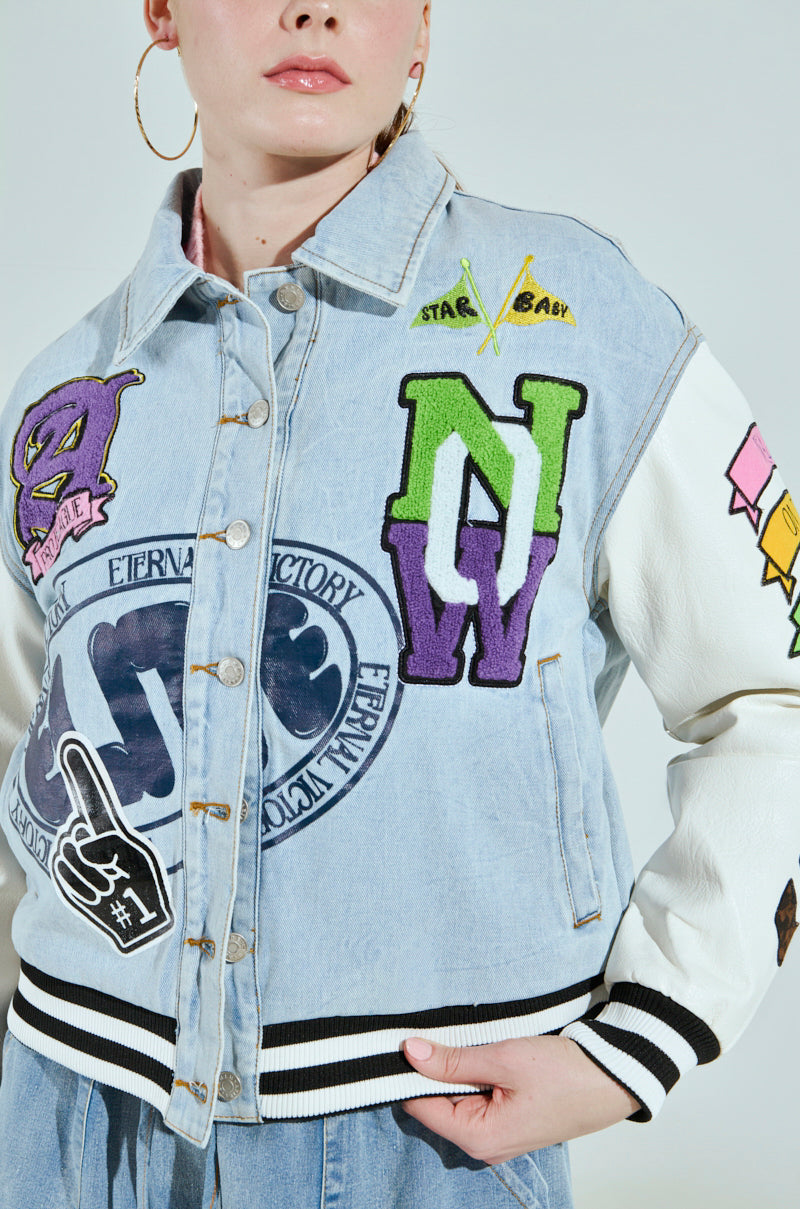 Dressed in an oversized ACADEMIA DENIM BOMBER adorned with vibrant patches and teamed with hoop earrings and a light shirt, the individual stands against a plain, neutral background.