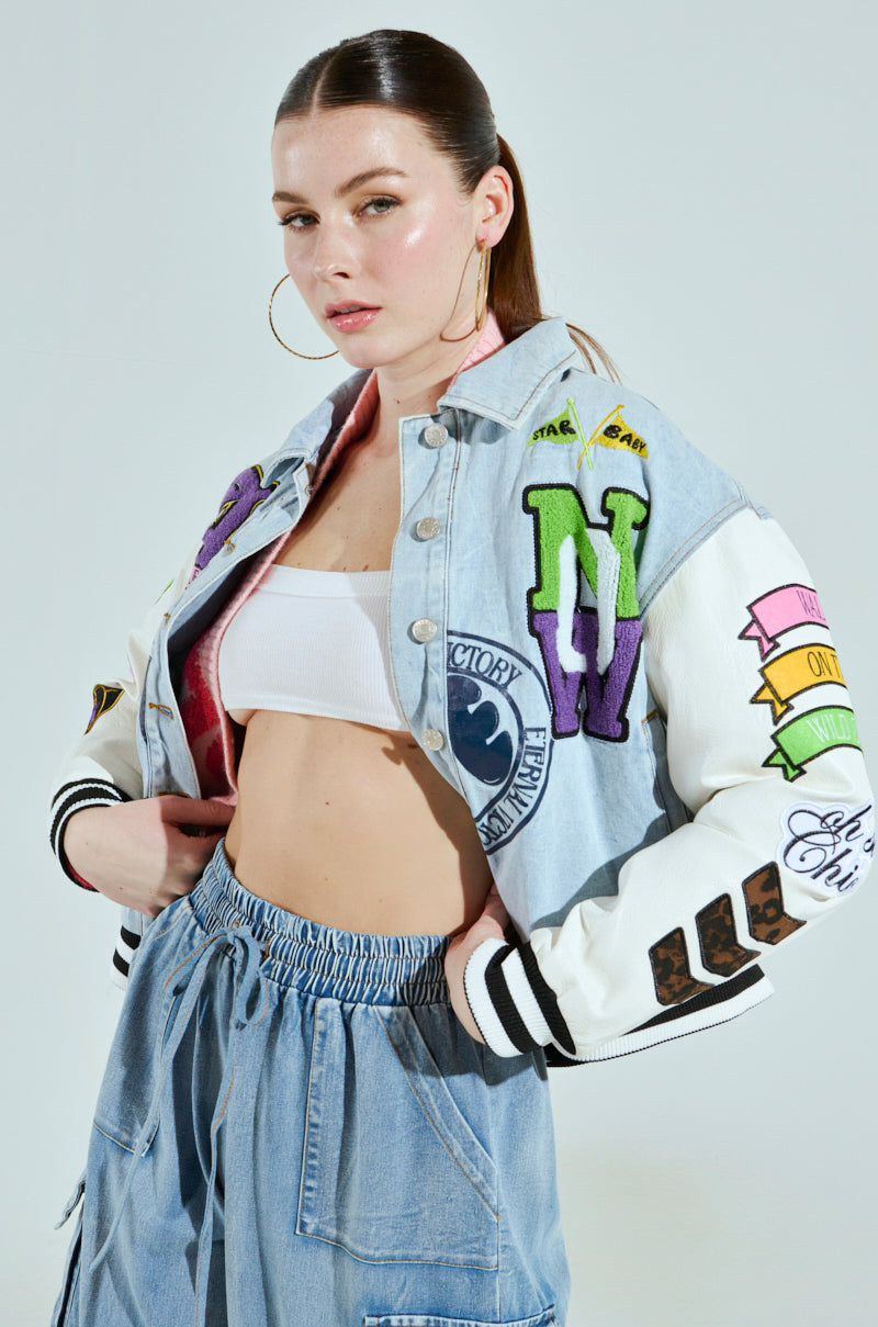 A person with long brown hair dons the vibrant ACADEMIA DENIM BOMBER, accessorized with large hoop earrings. The outfit features a white top and light denim pants, all set against a plain light gray background that highlights it beautifully.