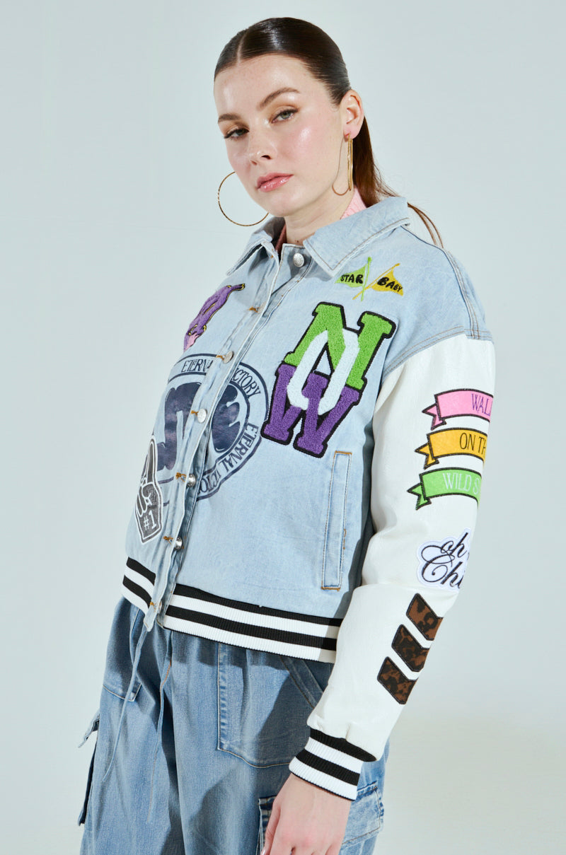 A person with long hair tied back is wearing the ACADEMIA DENIM BOMBER, an oversized jacket adorned with colorful patches, mixed media designs, large letters, and various motifs. They complete the look with denim pants and hoop earrings while gazing slightly to the side.