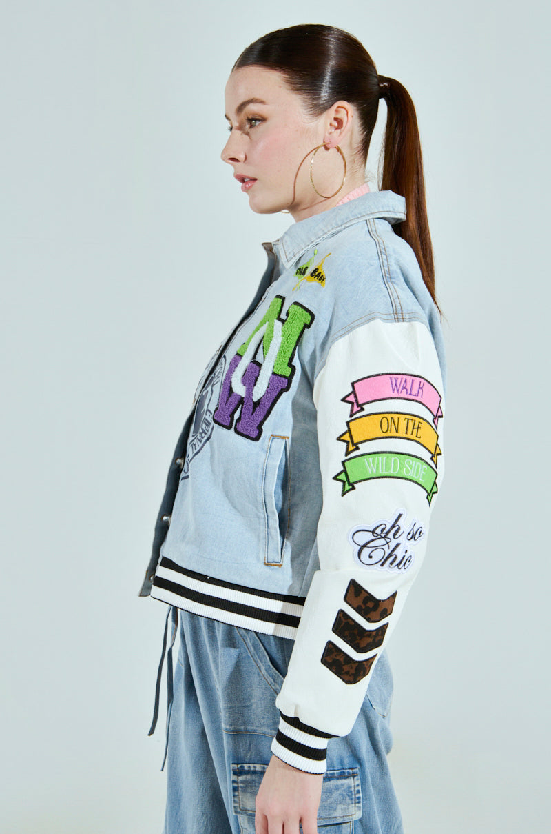 A woman with long dark hair in a ponytail, wearing large hoop earrings, is dressed in the ACADEMIA DENIM BOMBER jacket featuring colorful patches and "Walk on the Wild Side" text on the sleeve. She pairs it with matching denim pants as she stands in profile against a plain background.