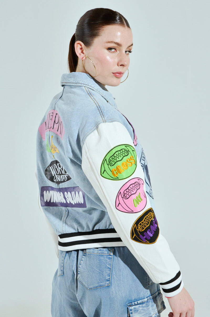 A person with brown hair tied back is wearing the ACADEMIA DENIM BOMBER, a light blue and white varsity jacket with colorful patches like lips and text. They pair it with denim jeans, hoop earrings, and glance over their shoulder.
