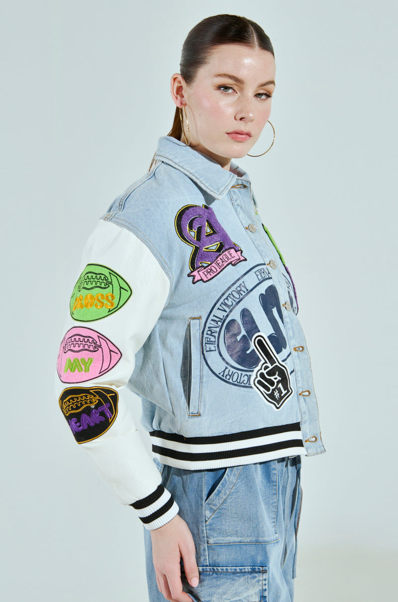 A person styled in the ACADEMIA DENIM BOMBER, an oversized light denim jacket with colorful patches and mixed media designs. It showcases letters and symbols on the sleeves and chest. Paired with light blue jeans, they have long hair tied back and large hoop earrings.