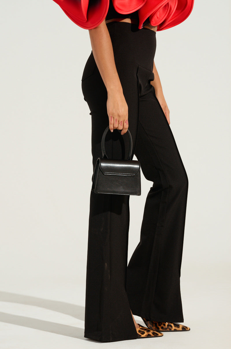 A person wearing the "Memories That Last Pant with Slit in Black" and leopard-print shoes is holding a small black handbag. Part of a red ruffled top is visible at the top of the image against a plain, light-colored background.