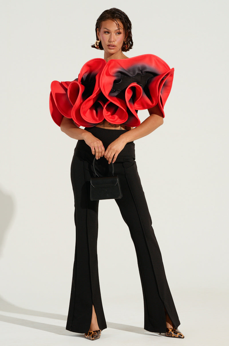 A person poses in an artistic outfit featuring a sculptural red and black ruffled top, the MEMORIES THAT LAST PANT WITH SLIT IN BLACK, and leopard print shoes. They hold a small black handbag against a plain background.