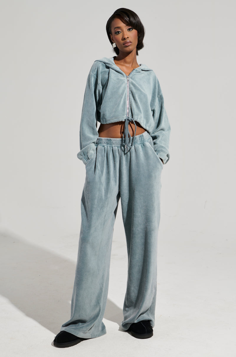 Dressed in the AKIRA Label attire, a person stands with hands in pockets, showcasing a light grey tracksuit that includes a zip-up cropped hoodie and high-rise FREYA SWEATPANTS with wide-leg design. The stylish black footwear completes the look against a plain white background.