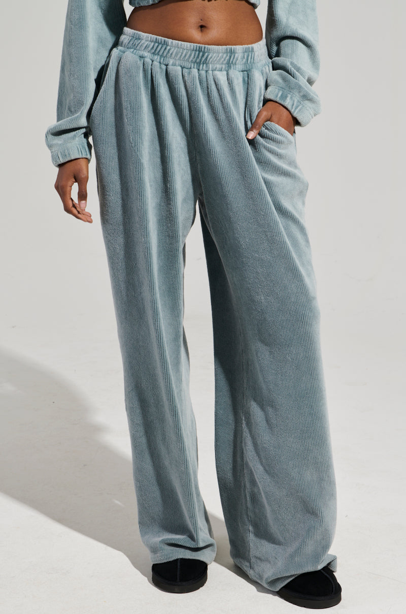 Wearing the FREYA SWEATPANTS in a light blue corduroy finish, featuring a loose fit and an elastic waistband, someone has their right hand tucked into one of the pockets. The sweatpants are stylishly combined with black shoes against a simple, light-colored background.