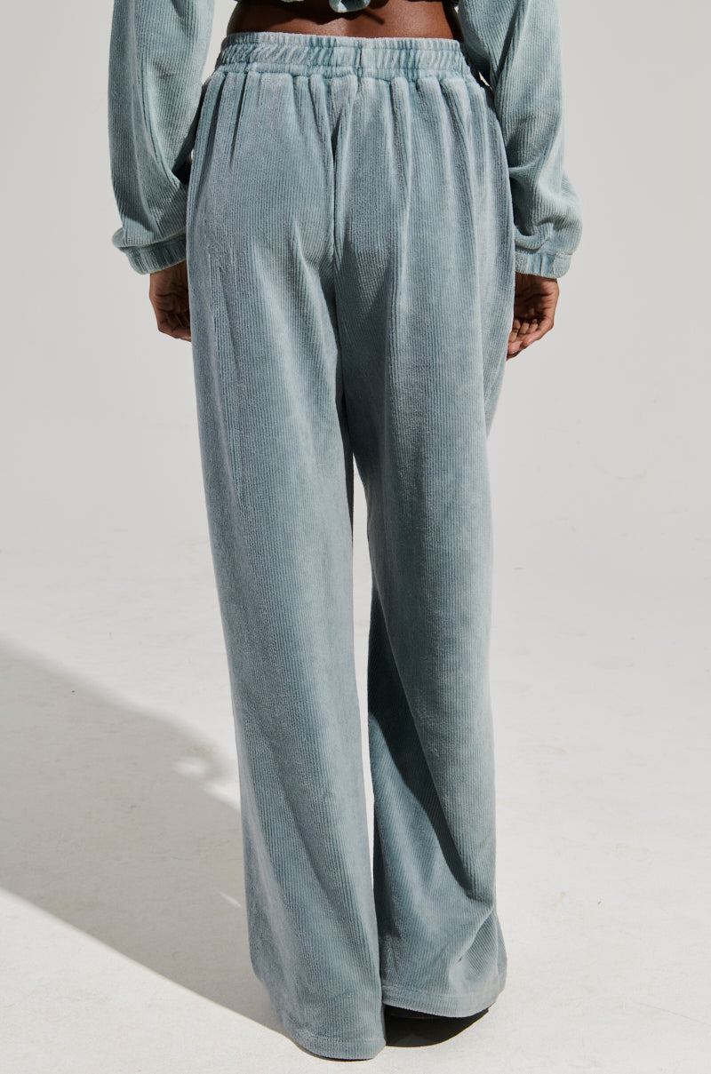 A person is seen from behind wearing the FREYA SWEATPANTS in a light blue-gray corduroy fabric paired with a matching top, highlighting the relaxed, wide-leg design similar to high-rise sweatpants. The simple light-colored backdrop enhances the AKIRA Label aesthetic.