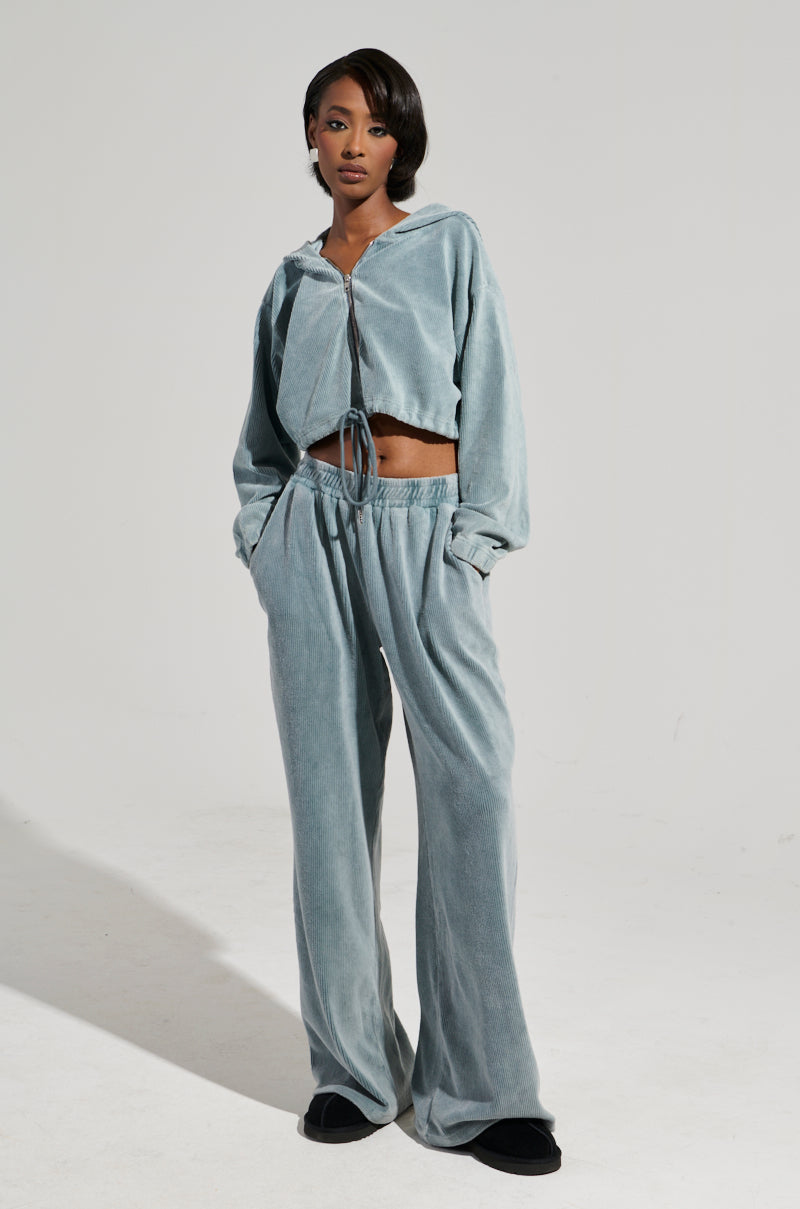 A person is standing in a light gray velour tracksuit and black slippers against a plain white background. Sporting the FREYA SWEATPANTS from the AKIRA Label, the ensemble includes a zip-up hoodie. The individual has short hair and poses confidently with hands in pockets.