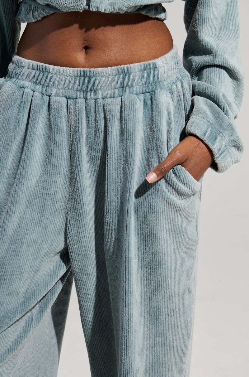 A person wearing the FREYA SWEATPANTS from AKIRA Label in a light green velour texture casually places their hand in the pocket. This look includes high-rise sweatpants with an elastic waistband paired with a cropped top, set against a plain, neutral background.