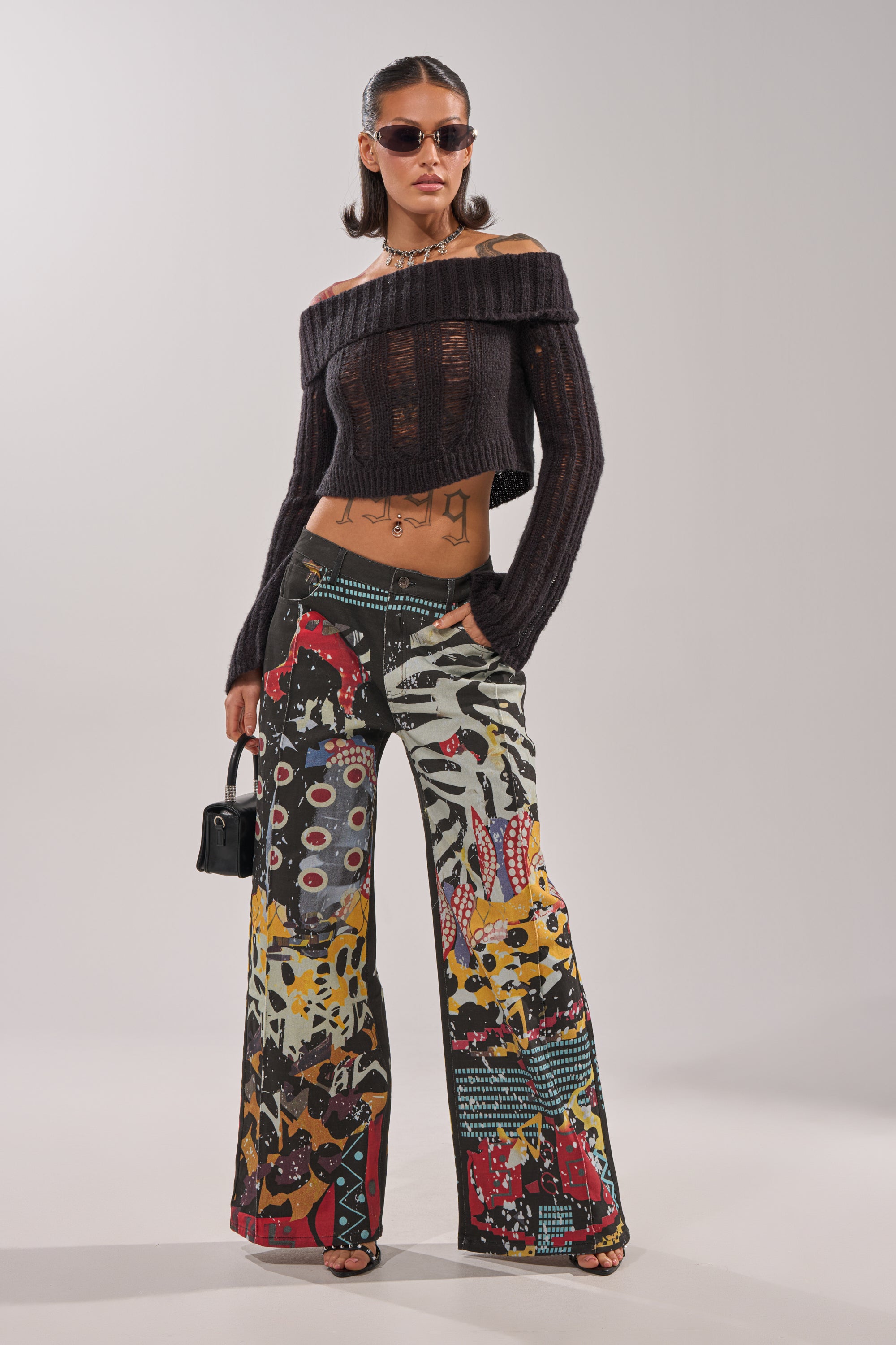 A woman stands confidently, wearing sunglasses, a black off-the-shoulder cropped sweater, and SOMETHING DIFFERENT PRINTED BLACK WASH JEANS with an abstract patchwork pattern, while holding a small black handbag. 1