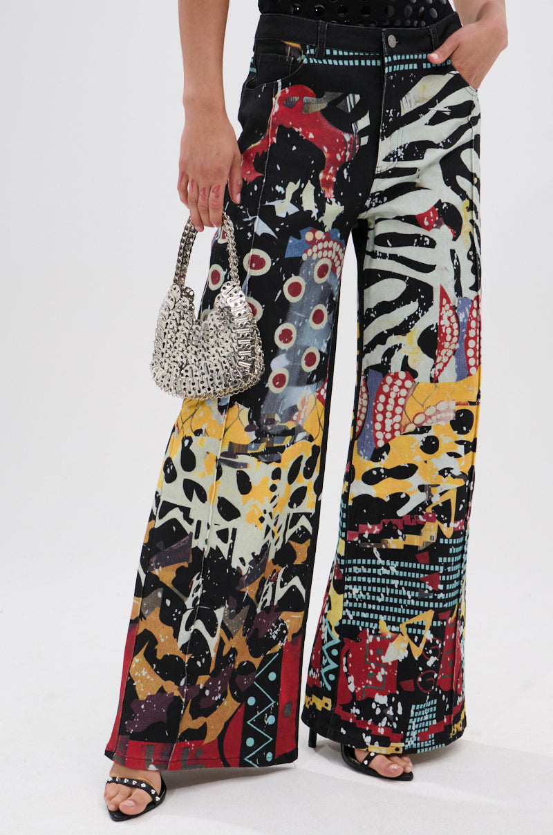 A person wears SOMETHING DIFFERENT PRINTED BLACK WASH JEANS with a bold abstract pattern, black sandals, and holds a small beaded handbag. The image is cropped at the torso to highlight the jeans and statement accessories.