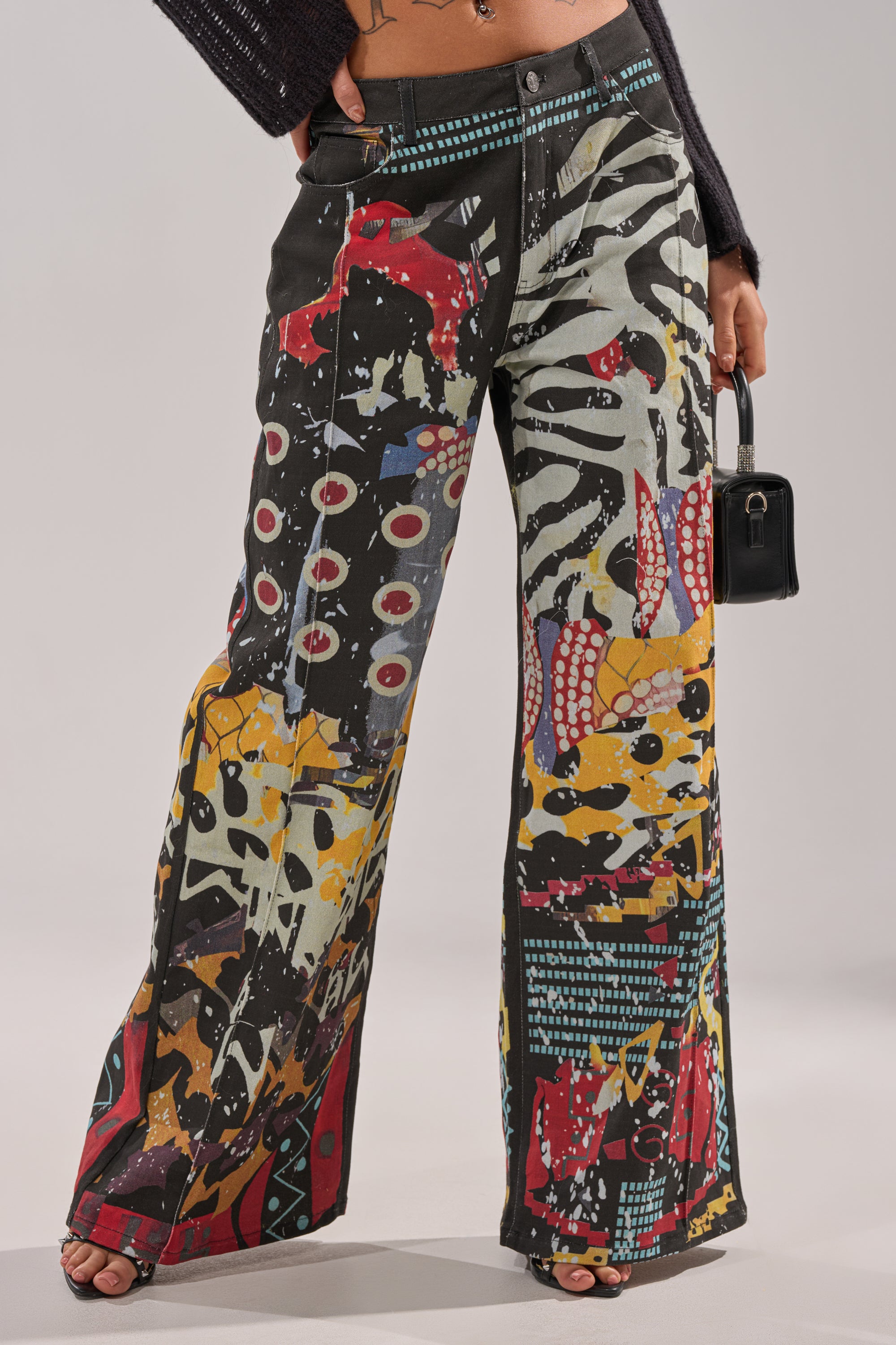 A person wearing SOMETHING DIFFERENT PRINTED BLACK WASH JEANS featuring a bold, abstract multicolored pattern, paired with a small black handbag. Only the lower torso and legs are visible. 2