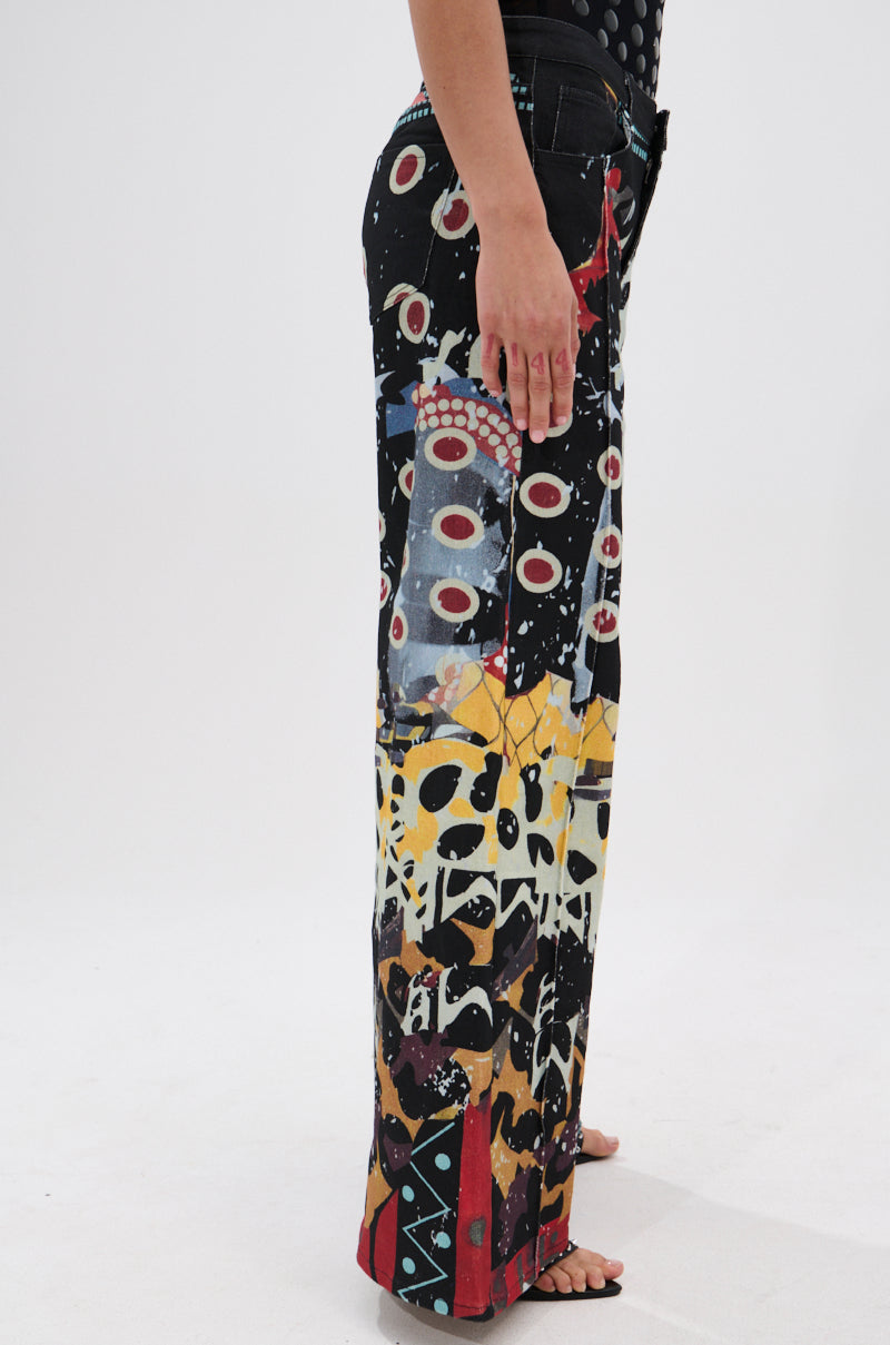 A person stands barefoot on a white background wearing SOMETHING DIFFERENT PRINTED BLACK WASH JEANS, which feature wide legs and a bold, colorful abstract pattern of circles, splashes, and geometric shapes; their arm rests by their side.