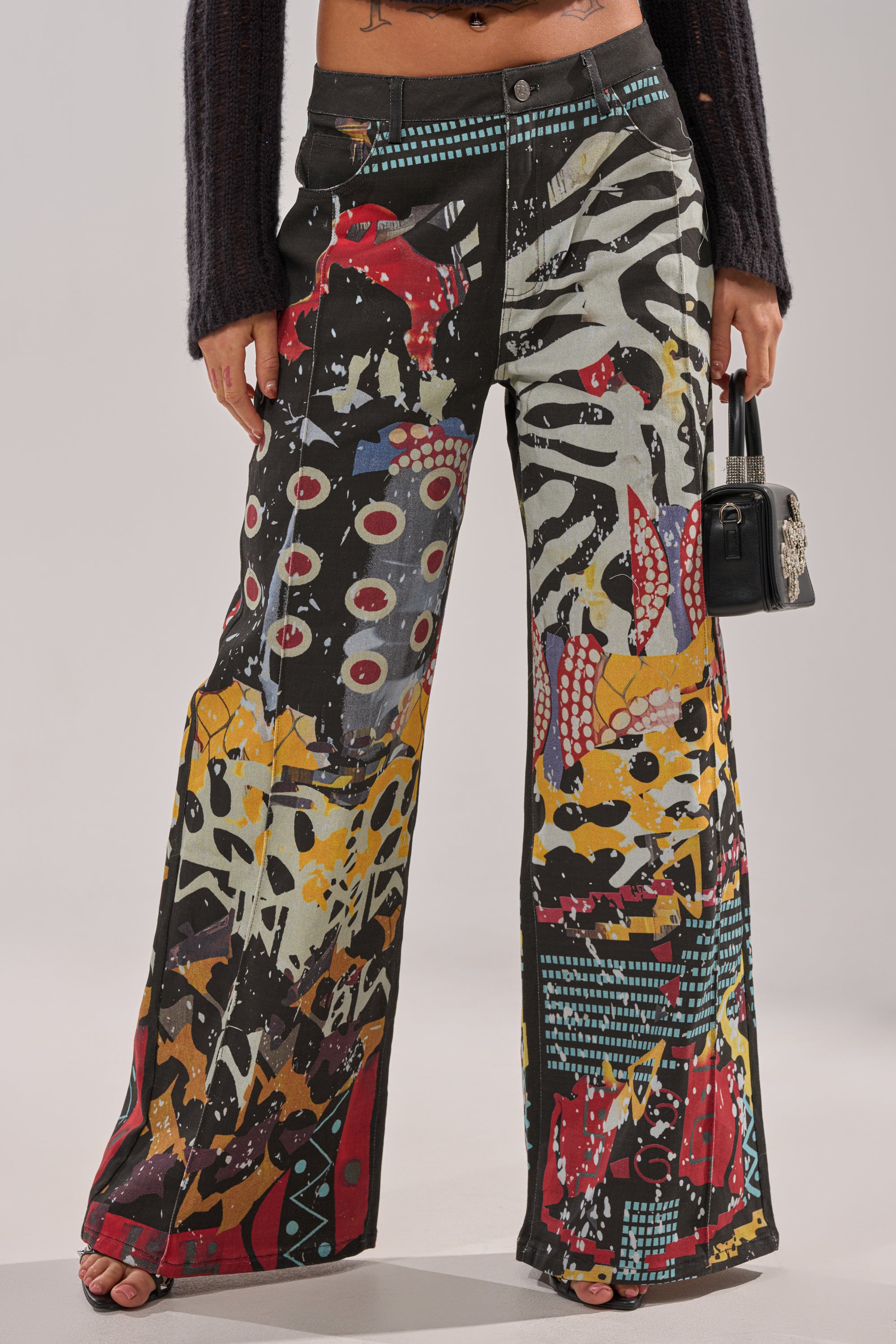 A person wears the SOMETHING DIFFERENT PRINTED BLACK WASH JEANS, which feature a bold, colorful abstract design with various shapes and prints, and holds a small black handbag. The upper body is partially visible. 0