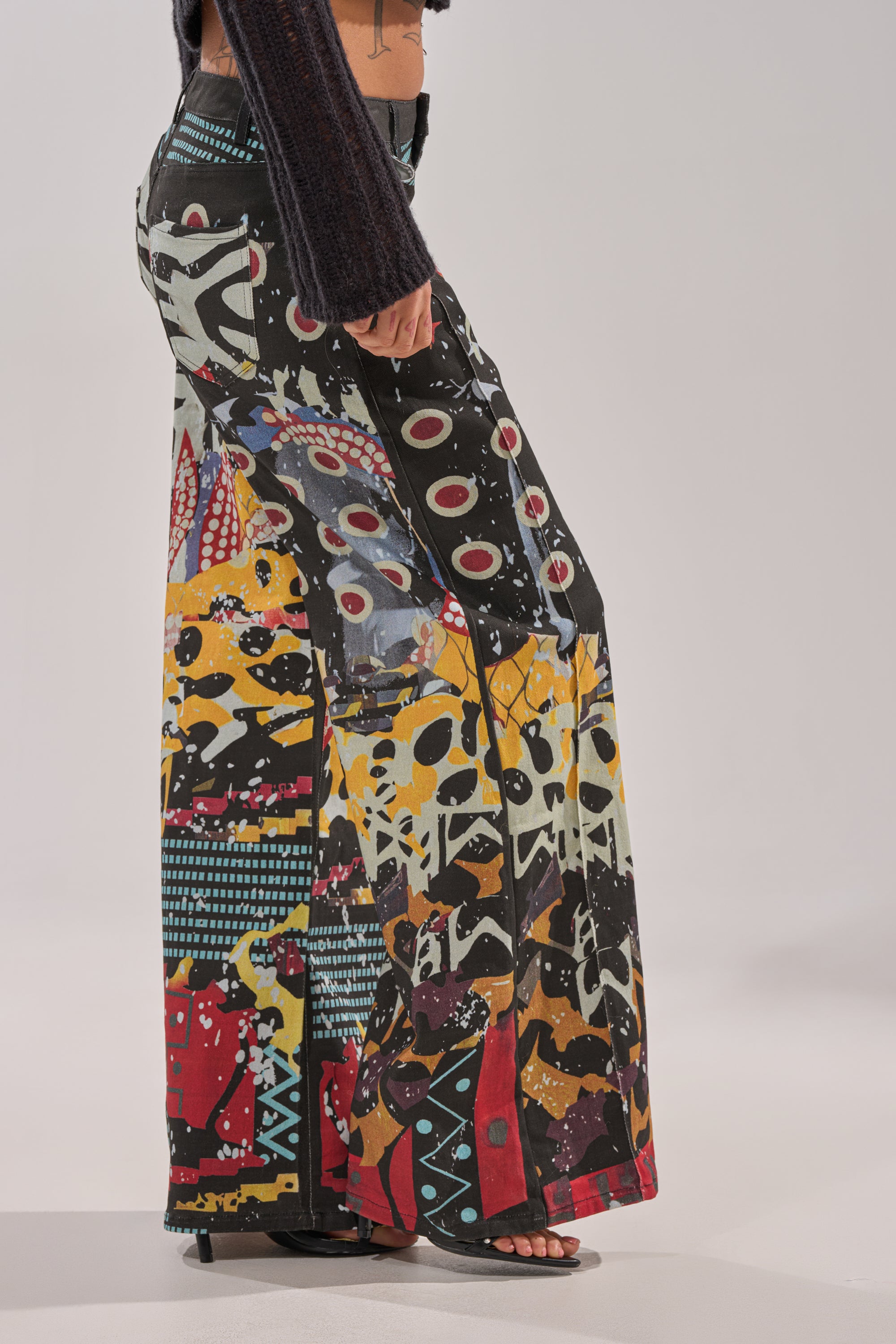 Someone wearing SOMETHING DIFFERENT PRINTED BLACK WASH JEANS featuring a bold, colorful abstract pattern, paired with black heeled sandals and a dark long-sleeve top, poses against a light background. 3