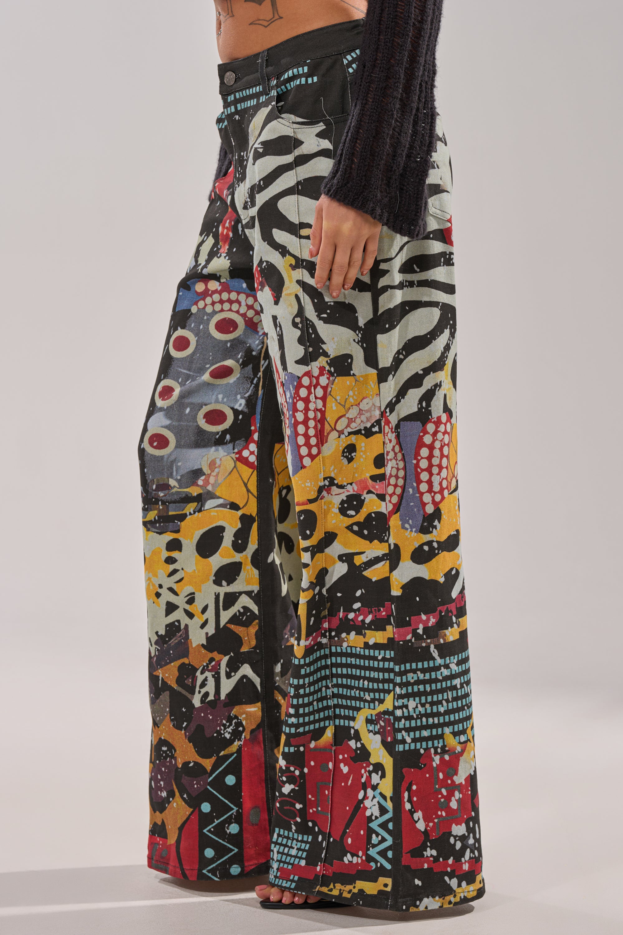 Someone wears SOMETHING DIFFERENT PRINTED BLACK WASH JEANS featuring a wide-leg fit and colorful, abstract graffiti patchwork in red, yellow, black, white, and blue. Their midriff and right hand are visible. 4