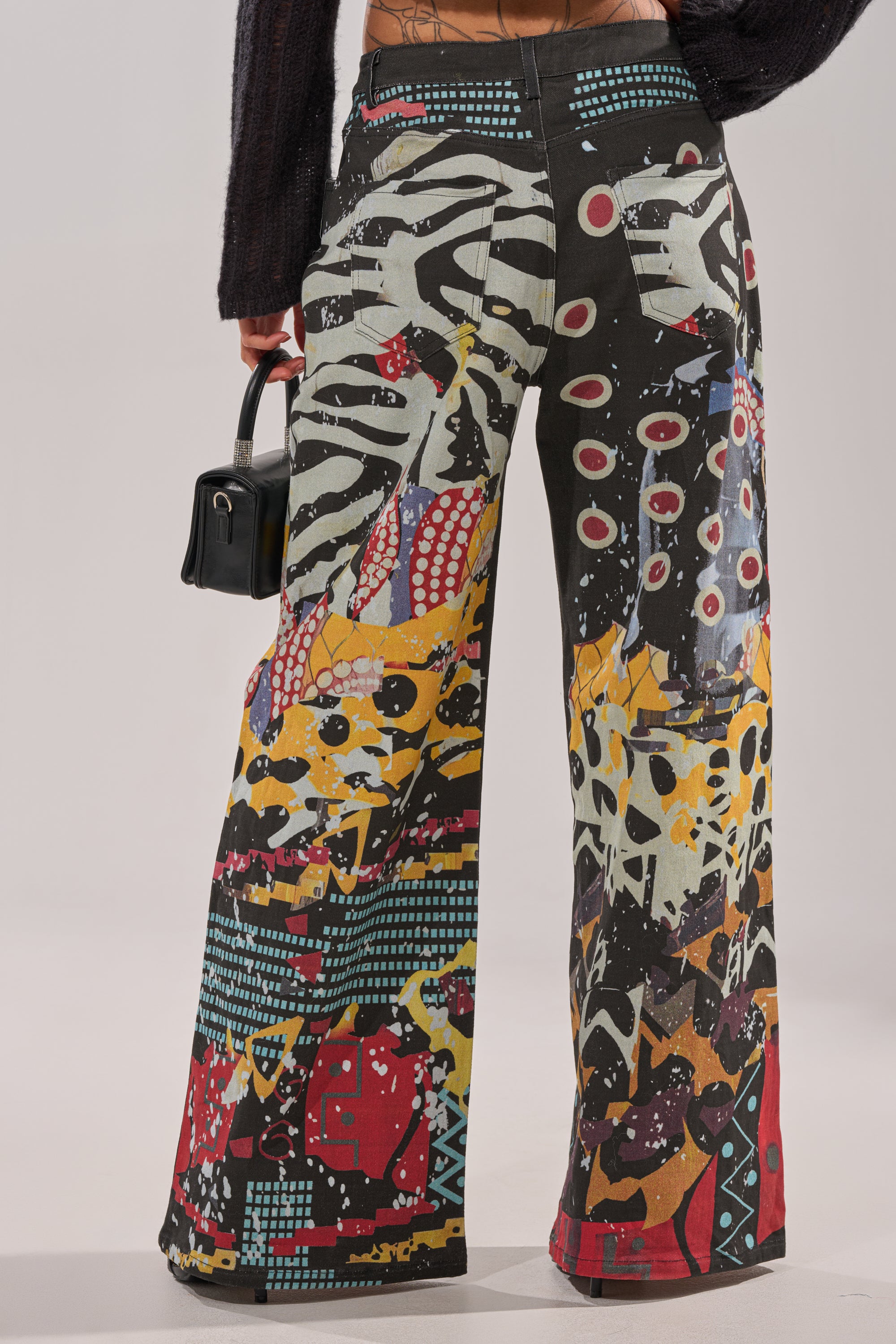 A person faces away from the camera, wearing SOMETHING DIFFERENT PRINTED BLACK WASH JEANS with red, yellow, blue, and white geometric and organic patterns, and holding a small black handbag. 5