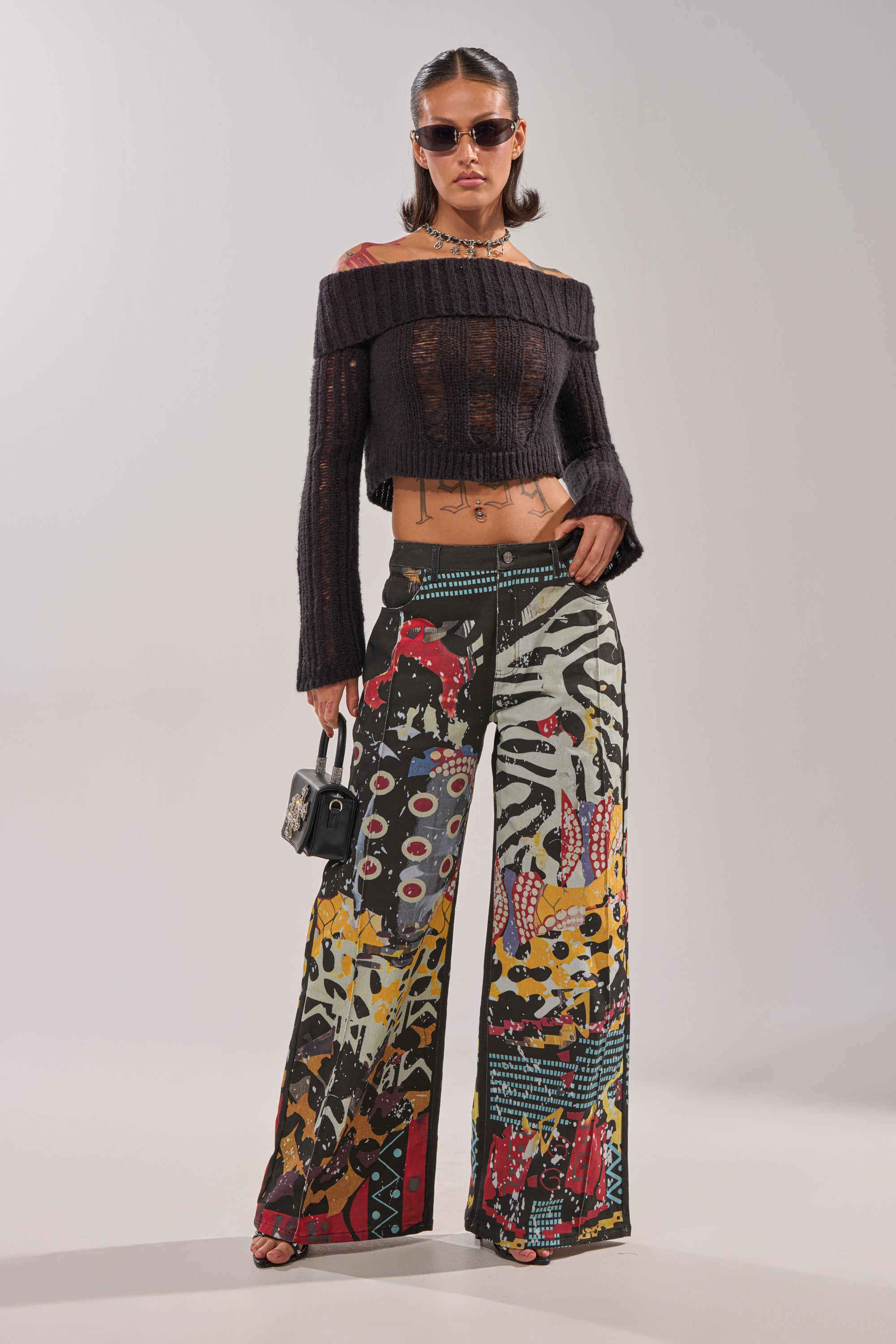 A woman stands against a plain light background wearing sunglasses, a black off-the-shoulder cropped sweater, and SOMETHING DIFFERENT PRINTED BLACK WASH JEANS, holding a small black purse. 6