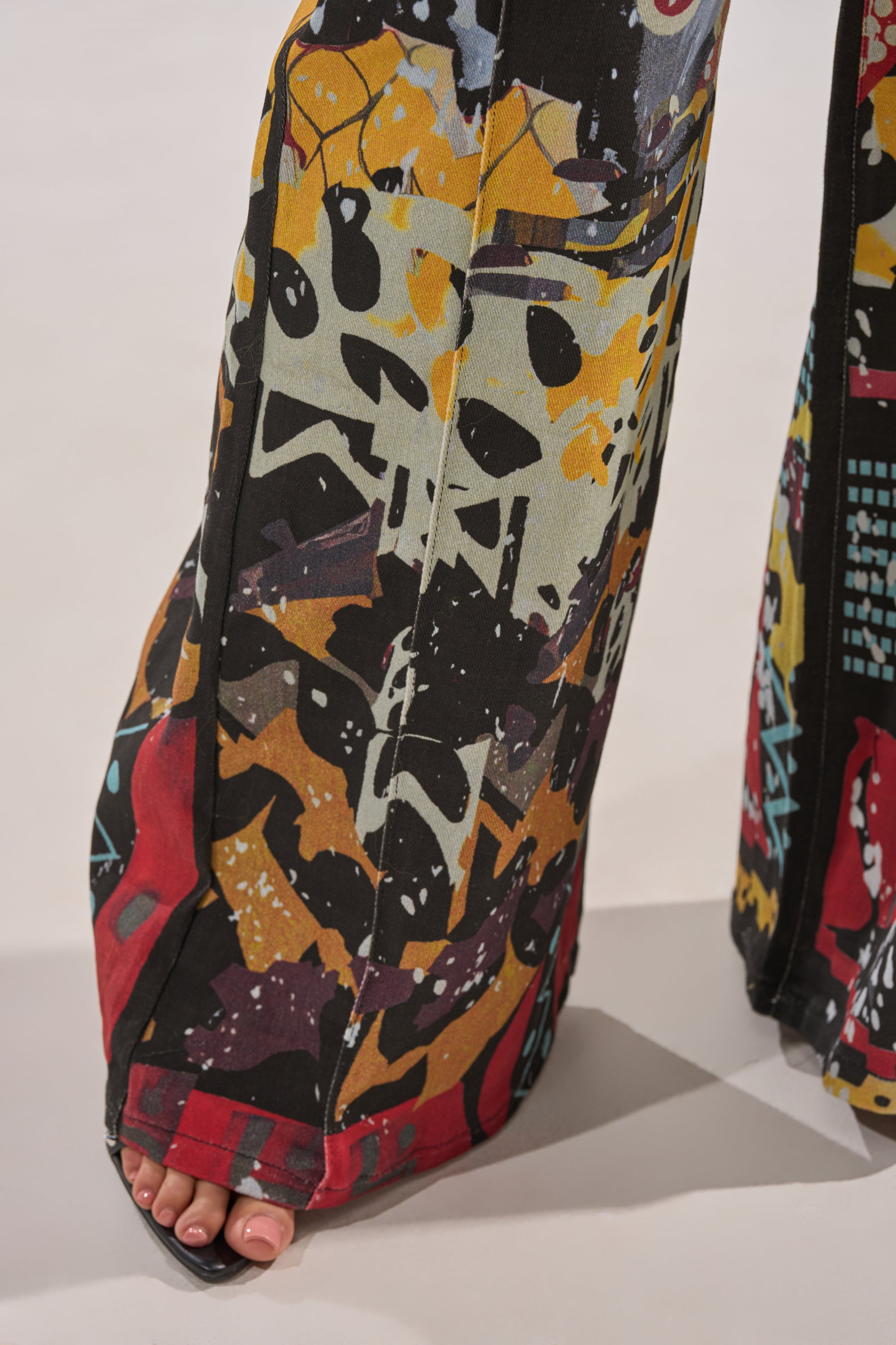 A close-up of a person wearing SOMETHING DIFFERENT PRINTED BLACK WASH JEANS featuring a bold graffiti-style abstract pattern in black, yellow, red, and blue. Bare toes peek out from beneath the wide-leg hem. 7