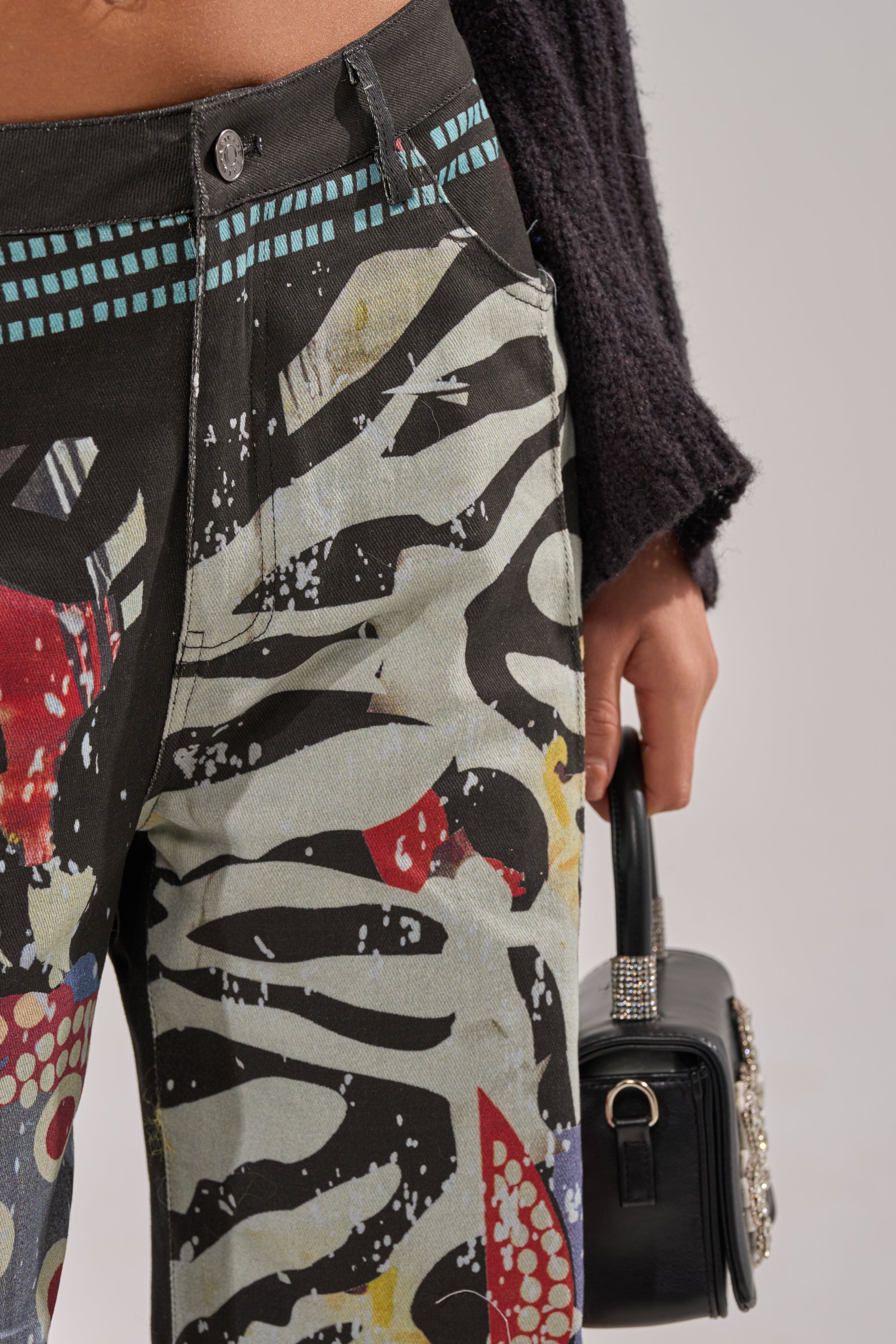 Close-up of a person wearing SOMETHING DIFFERENT PRINTED BLACK WASH JEANS with multicolor graffiti designs, holding a rhinestone-detailed black handbag, and dressed in a dark textured sweater. 8