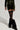 A person dressed in BRAT RHINESTONE VELVET SHORT—a dark green velvet jacket and shorts featuring rhinestone embellishments—enhanced by black knee-high socks and fluffy black boots, set against a plain, light-colored background.