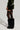 Facing sideways, the individual highlights the BRAT RHINESTONE VELVET SHORT in a rich dark green velvet, adorned with "Bratz" rhinestones. Complementing the outfit are black thigh-high stockings and oversized, furry black boots, all set against a simple light-colored backdrop.