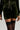 A person is styled in the BRAT RHINESTONE VELVET SHORT ensemble featuring a dark green velvet fabric and adorned with rhinestone embellishments, capturing Rockstar Dream vibes. The outfit consists of matching shorts and a top with a drawstring waist, complemented by black thigh-high socks against a simple light background.