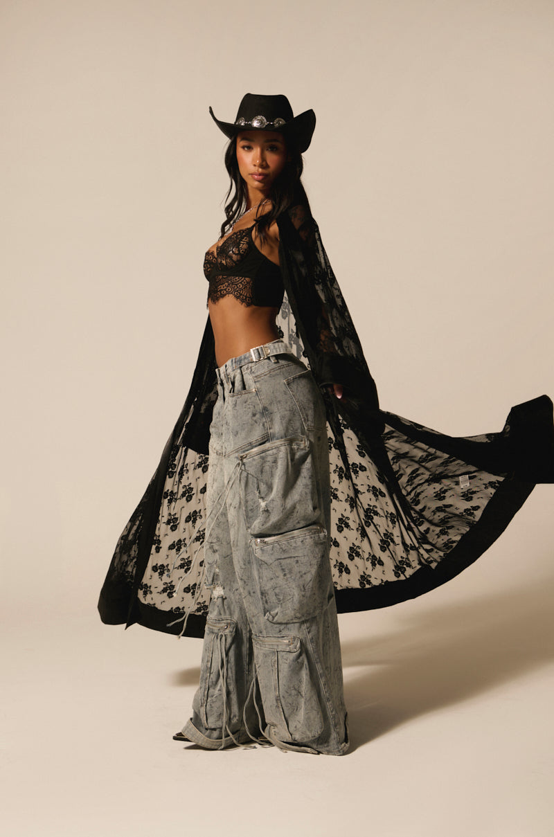 A woman poses confidently in a black cowboy hat, sheer lace top, and long lace robe. She's wearing PLACES TO BE RELAXED CARGO DENIM JEANS with multiple pockets and distressed detailing, standing out against a plain, neutral background.