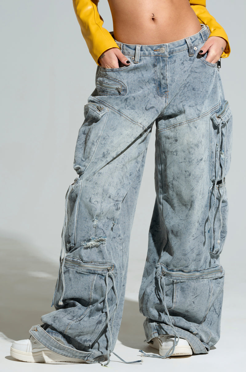A person is wearing the "PLACES TO BE RELAXED CARGO DENIM JEANS," featuring a baggy fit, multiple pockets, and distressed detailing with drawstrings and a mid-rise design. Paired with a yellow cropped top, these jeans boast a worn, faded appearance and are complemented by white sneakers to complete the outfit.