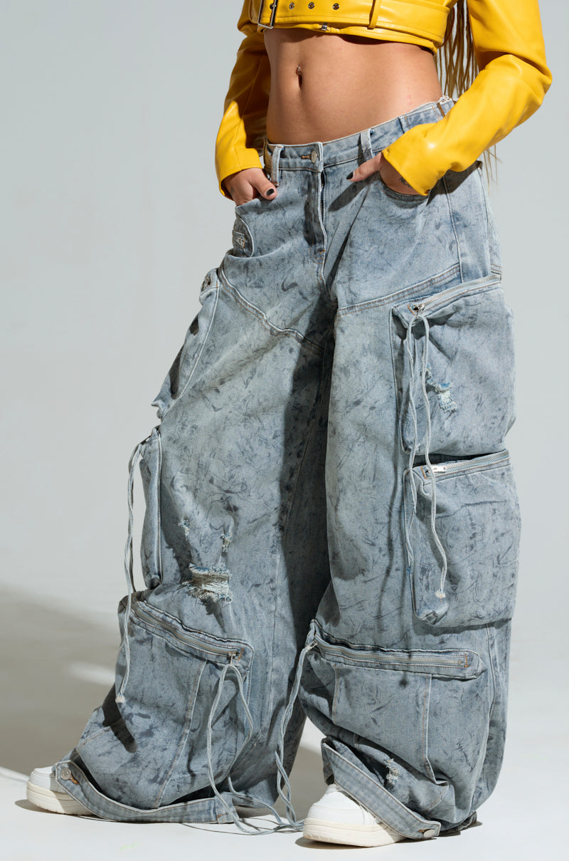Dressed in the PLACES TO BE RELAXED CARGO DENIM JEANS, featuring an oversized fit and light denim wash with a mid-rise design and multiple pockets, a person casually stands against a plain background. They've paired these stylish jeans with a yellow cropped jacket, their hands tucked into the roomy pockets. To complete this trendy look, white sneakers are visible at the bottom.