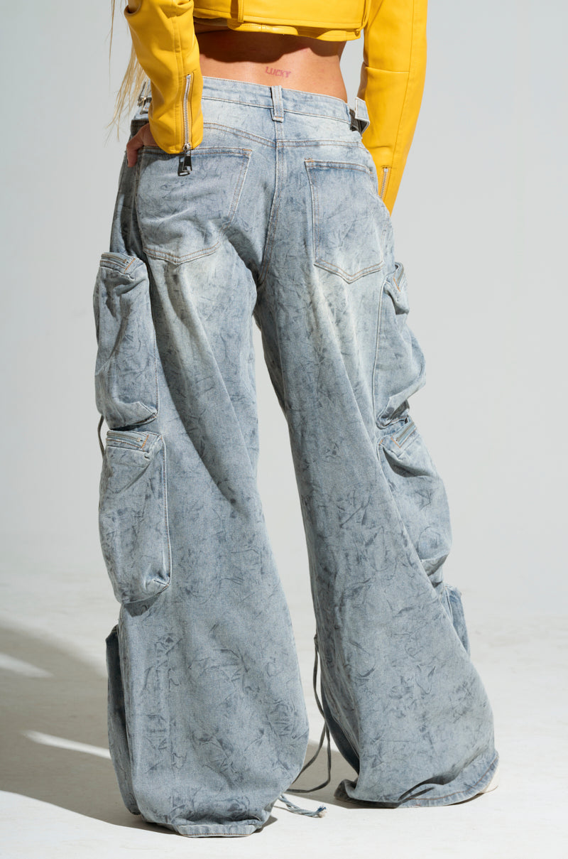 A person is wearing the PLACES TO BE RELAXED cargo denim jeans, which are light blue and oversized with multiple pockets. The jeans feature a mid-rise design, a loose, wide-leg fit with a marble wash, and distressed ripped detailing that adds flair. They perfectly complement the visible back and waist area for a stylish look when paired with a yellow jacket.