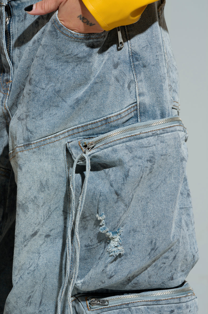 The image displays a detailed view of the PLACES TO BE RELAXED CARGO DENIM JEANS in a light blue, heavily washed finish. These jeans include multiple zippers and a large pocket with one featuring distressed ripped detailing and a frayed hole. A person is wearing these jeans alongside showing off their yellow sleeve, with "NOW" tattooed on their hand, enhancing the relaxed silhouette.