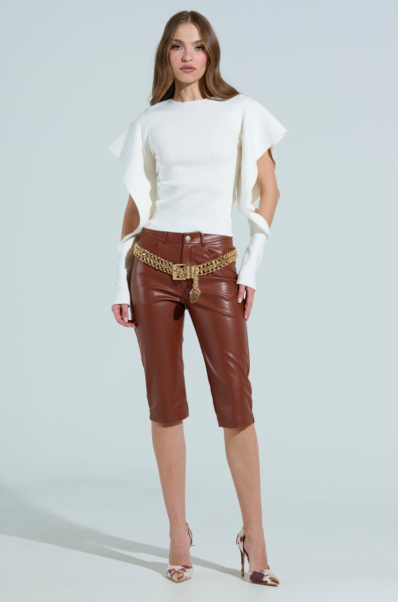 A person stands against a plain background wearing a white top with unique sleeve cutouts, SAMANTHA faux leather capri pants in brown, a chunky gold chain belt, and high-heeled shoes in a multicolored pattern.