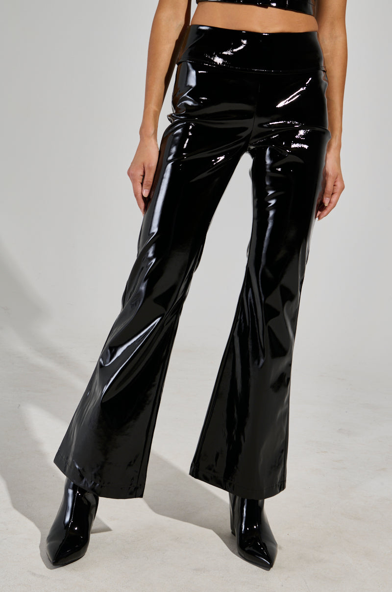 A person dressed in the SEXY FOX PANT in shiny black patent vinyl, complemented by matching glossy heeled boots. The fit and flare design beautifully reflects light, offering a sleek and stylish look, while the plain background accentuates their fashion statement.