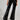A person dressed in the SEXY FOX PANT in shiny black patent vinyl, complemented by matching glossy heeled boots. The fit and flare design beautifully reflects light, offering a sleek and stylish look, while the plain background accentuates their fashion statement.