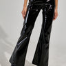 A person dressed in the SEXY FOX PANT in shiny black patent vinyl, complemented by matching glossy heeled boots. The fit and flare design beautifully reflects light, offering a sleek and stylish look, while the plain background accentuates their fashion statement.