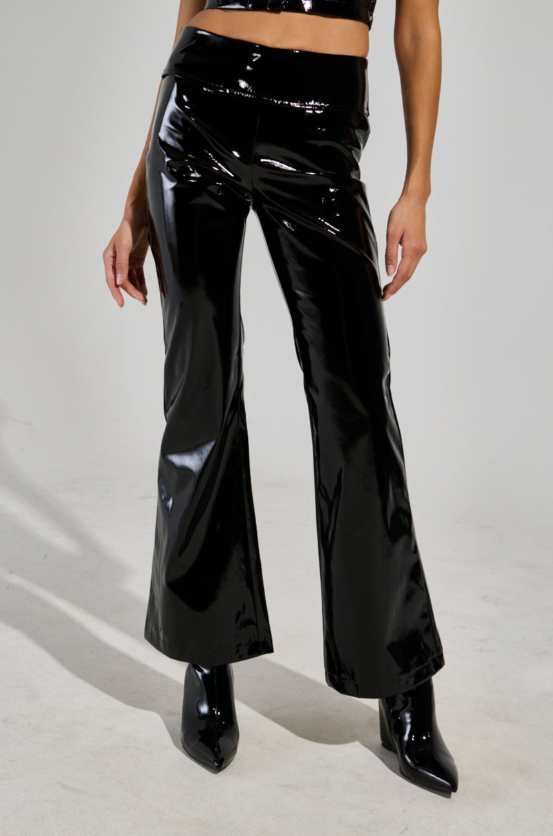 A person dressed in the sleek, high-waisted SEXY FOX PANT, featuring a fit n’ flare silhouette and matched with heeled boots, poses confidently with one hand on their hip. The shiny black patent vinyl of the pants gleams effortlessly against a plain, neutral backdrop.