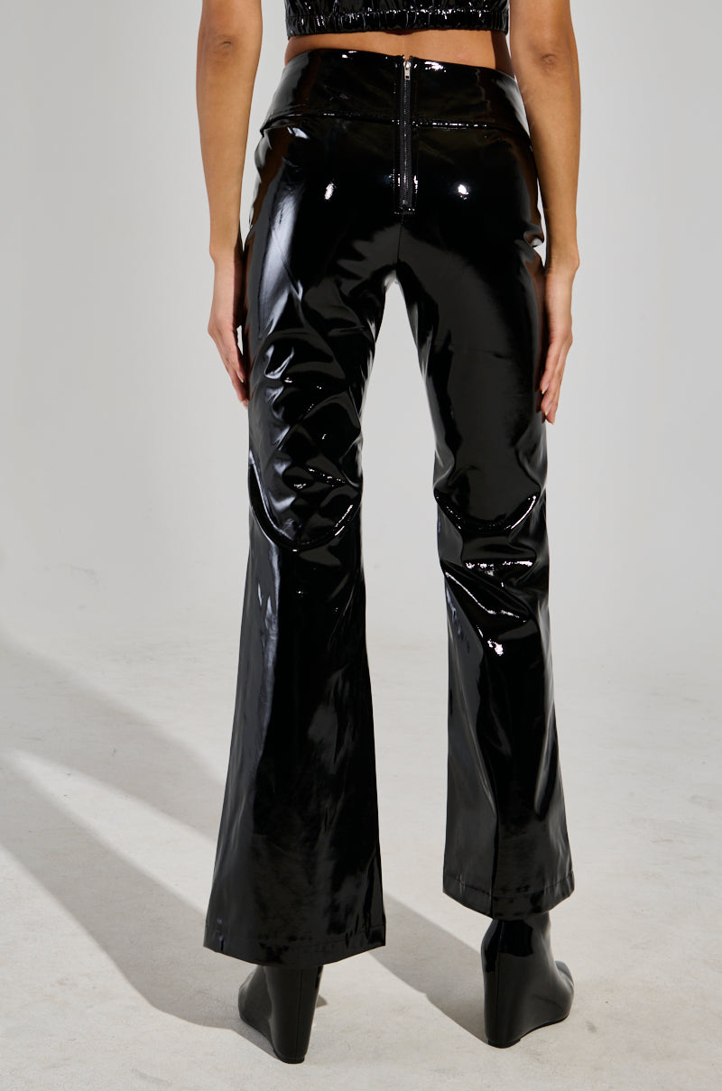 The person stands with their back turned, wearing the SEXY FOX PANT crafted from lustrous patent vinyl and featuring a fit n’ flare silhouette. These high-waisted pants elegantly flow into matching black boots, set against a plain, light-colored background that highlights the bold outfit.