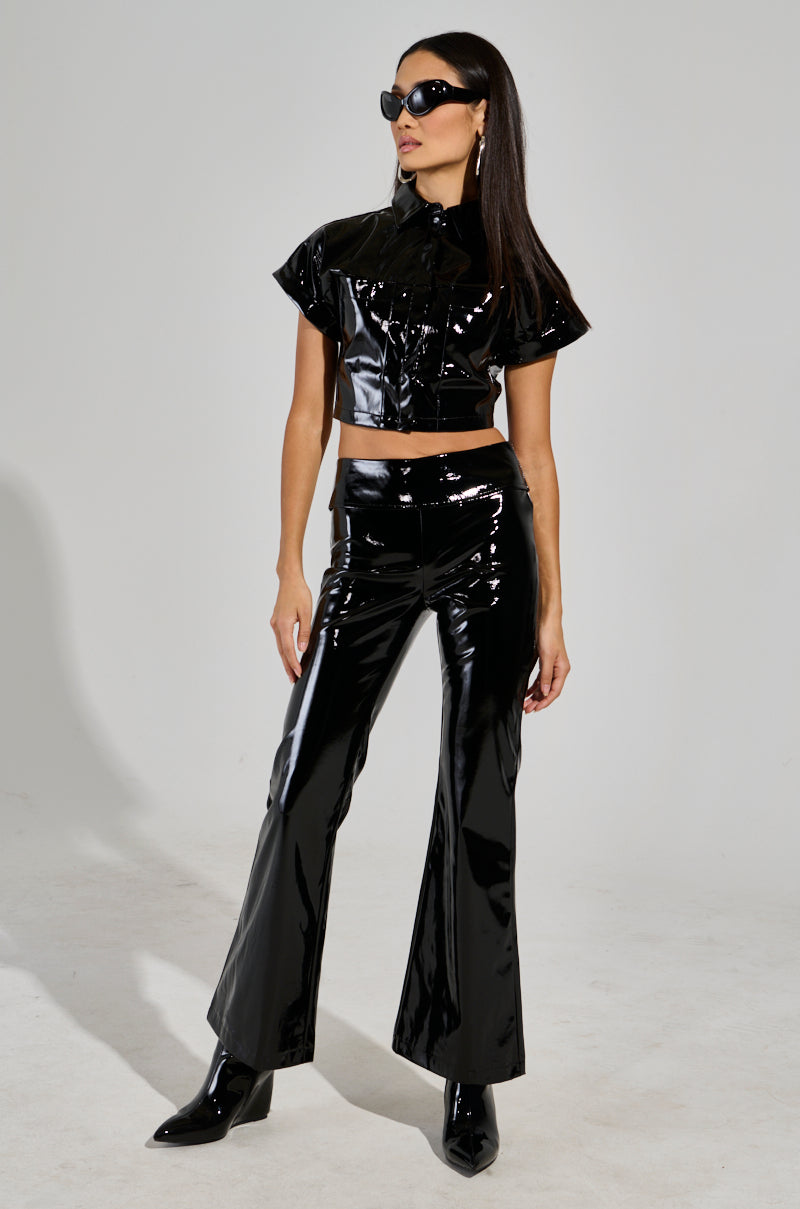 A woman models the Sexy Fox Pant ensemble against a plain background, wearing a sleek patent vinyl outfit that includes a short-sleeved crop top with a fit n' flare silhouette. She has long dark hair, dons sunglasses, and completes her look with black pointed boots.