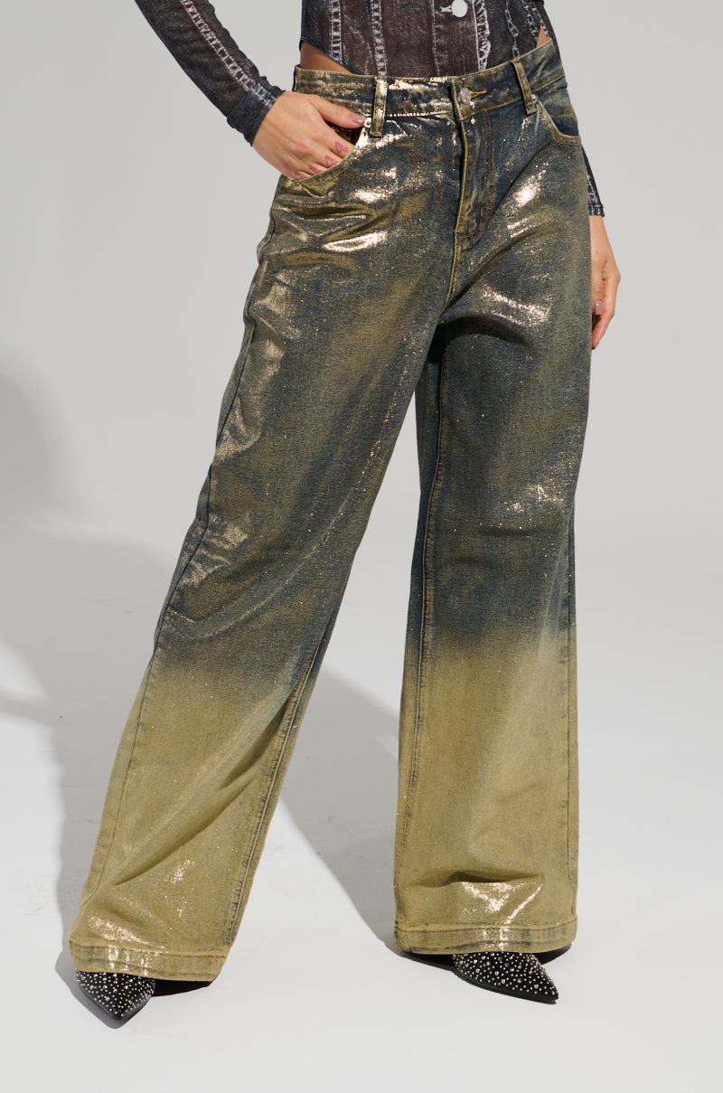 An individual showcases the ALL ABOUT THAT SHINE PANT, featuring high-rise denim in metallic gold with a gradient effect and a wide-leg style, complete with a shiny, glittery finish. These pants are ideally paired with pointed black shoes against a plain, light-colored backdrop.