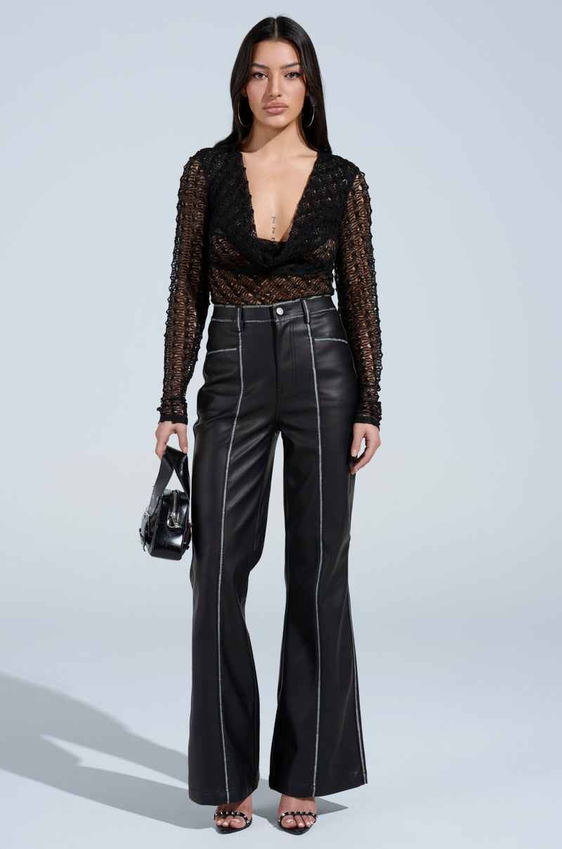 A woman poses in FLY GIRL FAUX LEATHER PANT—high-rise, flared leg with white seams—paired with a sheer black top, black heeled sandals, and a small black handbag against a light background. 4